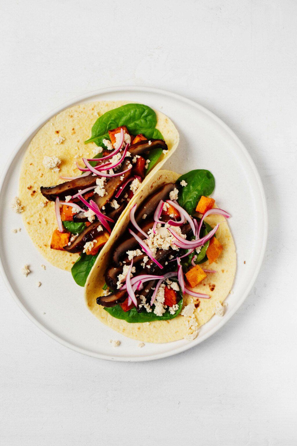 An overhead image of two neatly folded vegan portobello mushroom tacos, which are garnished with thinly sliced pickled red onion and plant-based "feta" cheese.