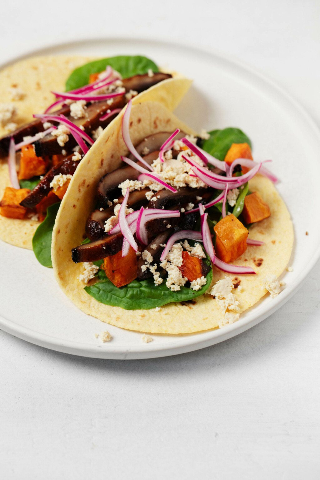 A round white plate holds vegan sweet potato and portobello mushroom tacos, each garnished with crumbled plant-based cheese and pickled onions.