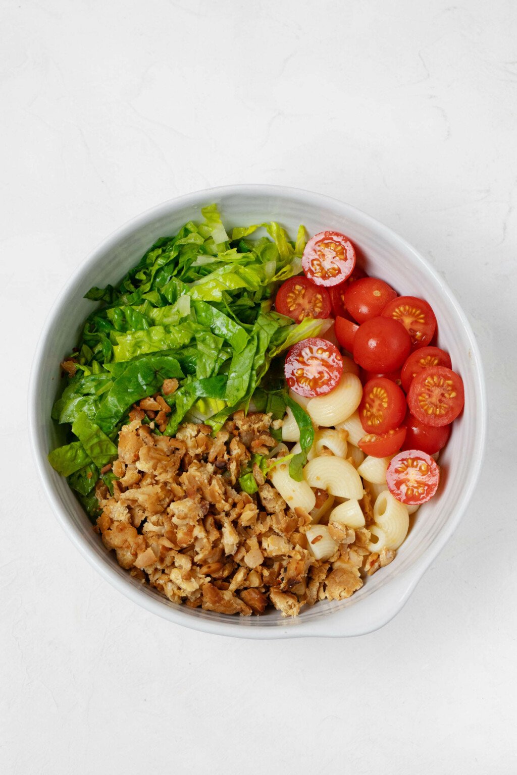 A large, round white mixing bowl is filled with tomatoes, tempeh bacon, cooked pasta, and chopped romaine lettuce.