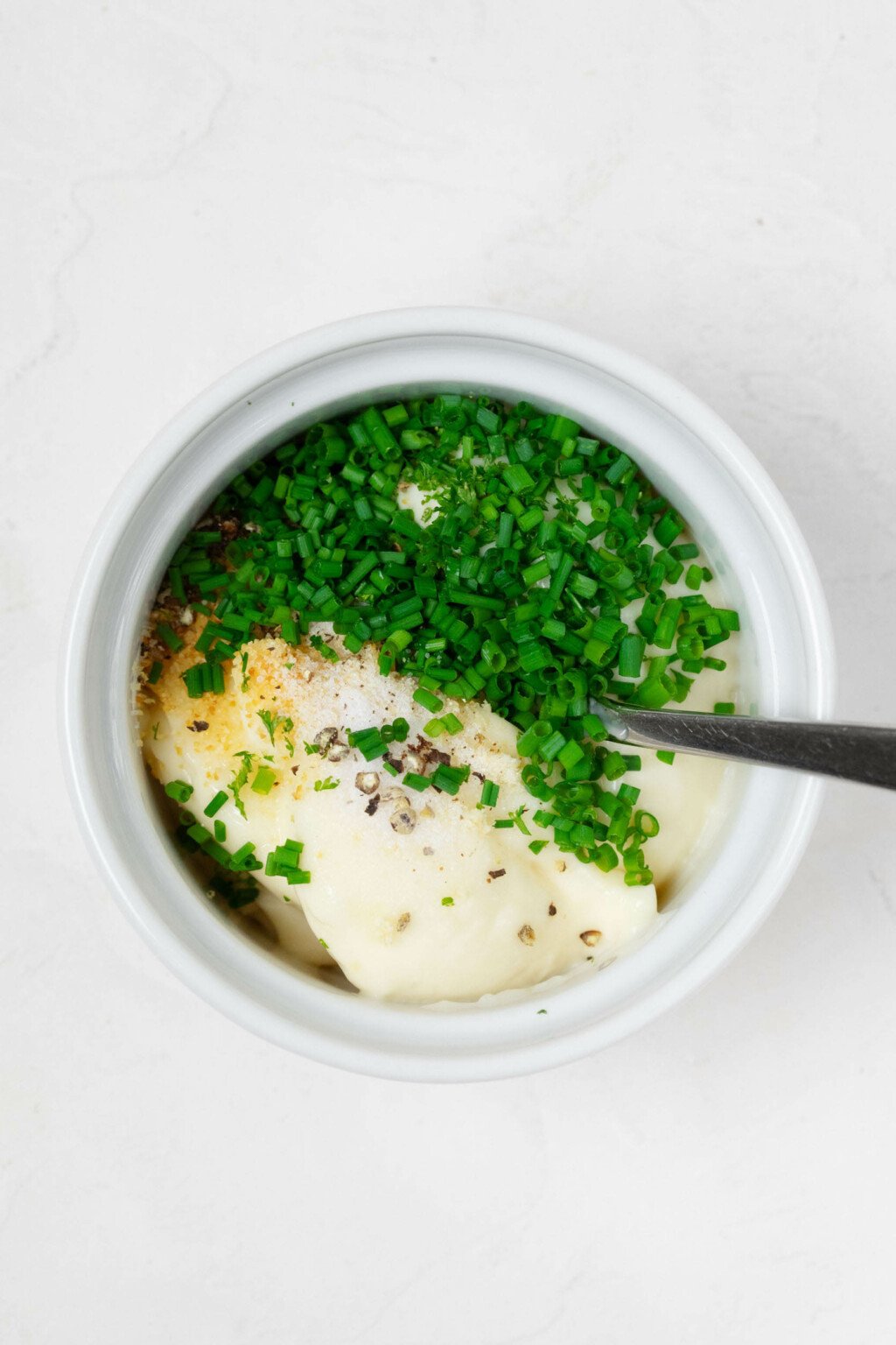 A small, white ramekin is filled with pale yellow mayonnaise and chopped green chives.