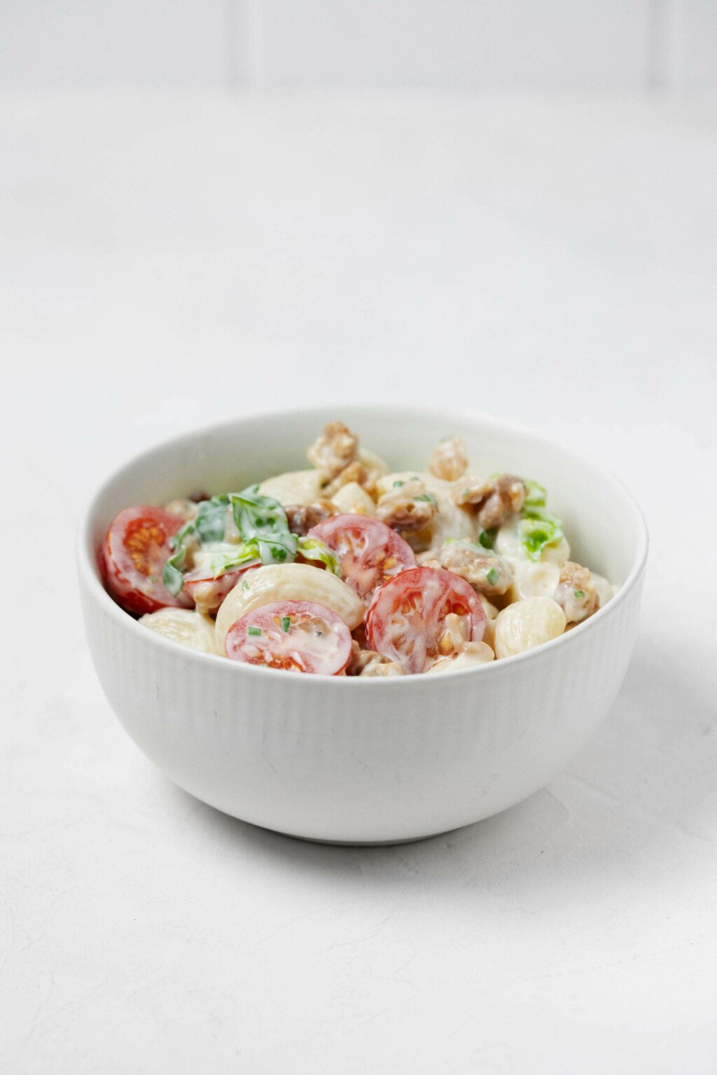 A round, white bowl is filled with a vegan BLT-inspired, creamy pasta salad. It rests on a white surface.