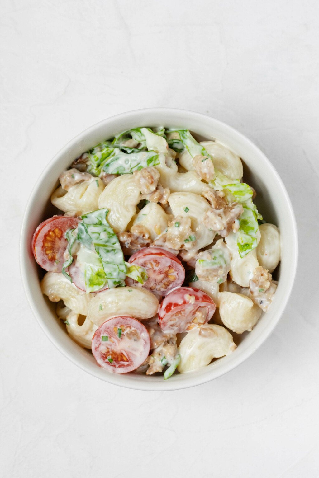 A white, round bowl is resting on a white surface. It holds the ingredients for a creamy, plant-based pasta salad.