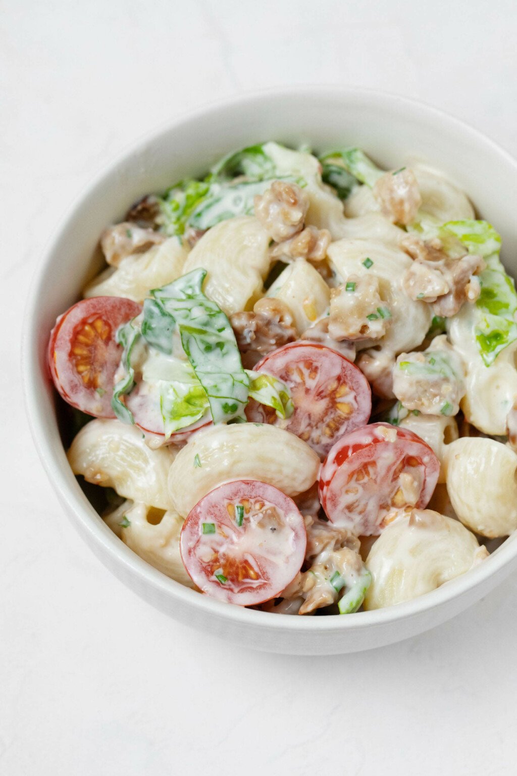 A round, white bowl is filled with a vegan BLT-inspired pasta salad, made with creamy dressing, cherry tomatoes, and chopped greens.