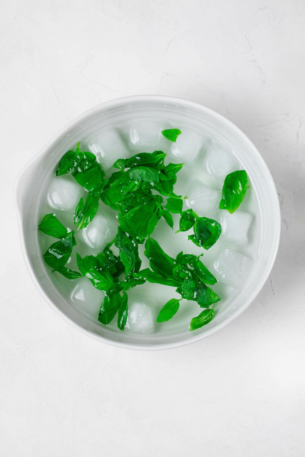 A large, white bowl of ice is flecked with basil leaves, which have been blanched and are cooling off.