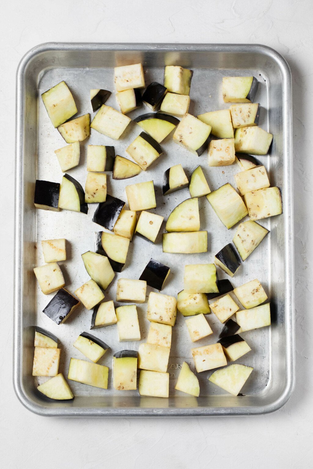 A silver baking sheet is topped with cubed eggplant, ready to be roasted.