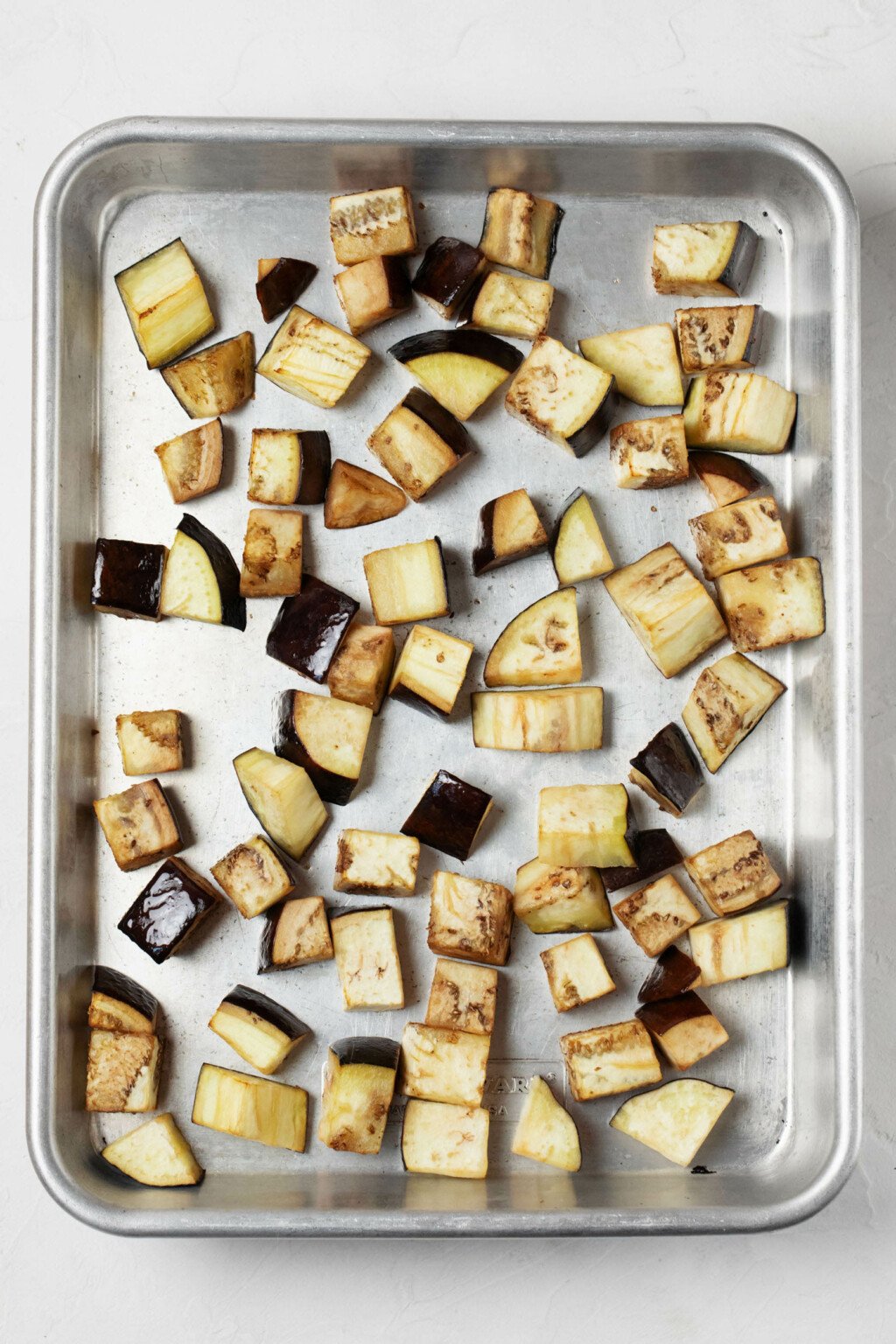 Roasted eggplant cubes are pictured on a rimmed, silver baking sheet.