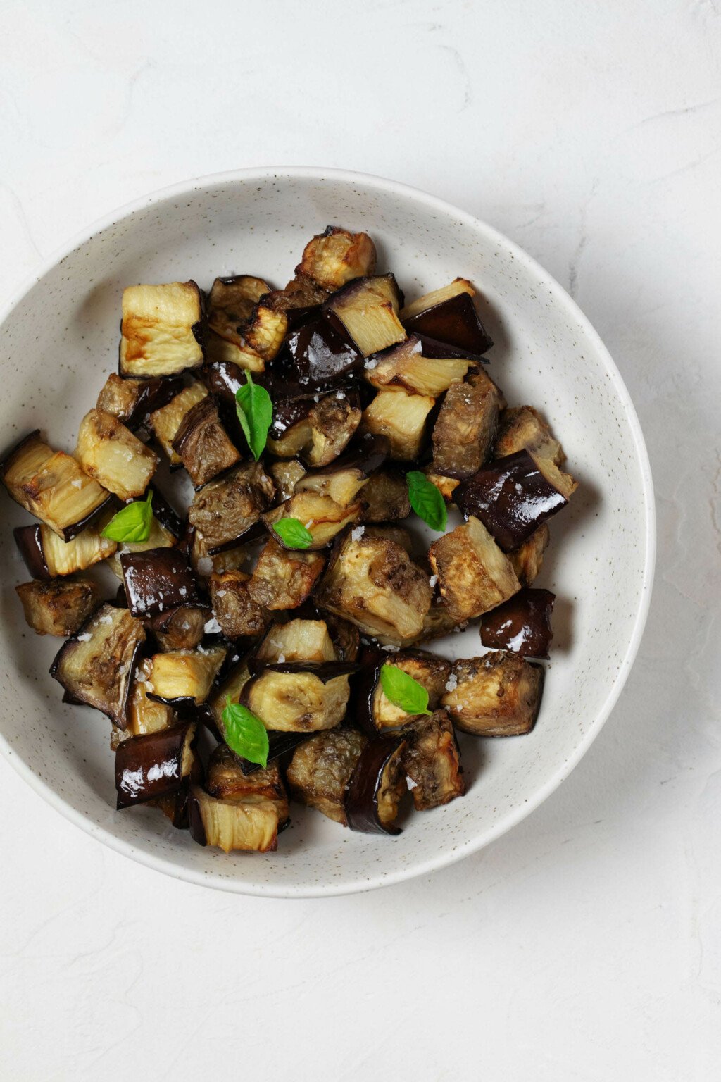 A white, ceramic bowl is filled with roasted vegetables and tiny leaves of fresh, green basil. 