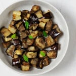 A white, ceramic bowl is filled with roasted vegetables and tiny leaves of fresh, green basil.