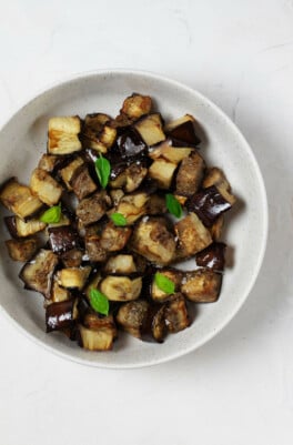 A white, ceramic bowl is filled with roasted vegetables and tiny leaves of fresh, green basil.