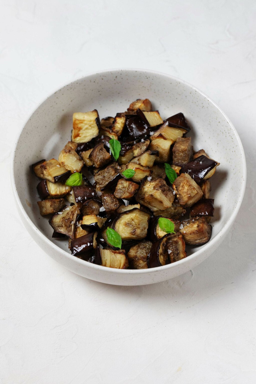 A white, ceramic bowl is filled with oven roasted eggplant cubes and tiny leaves of fresh, green basil.