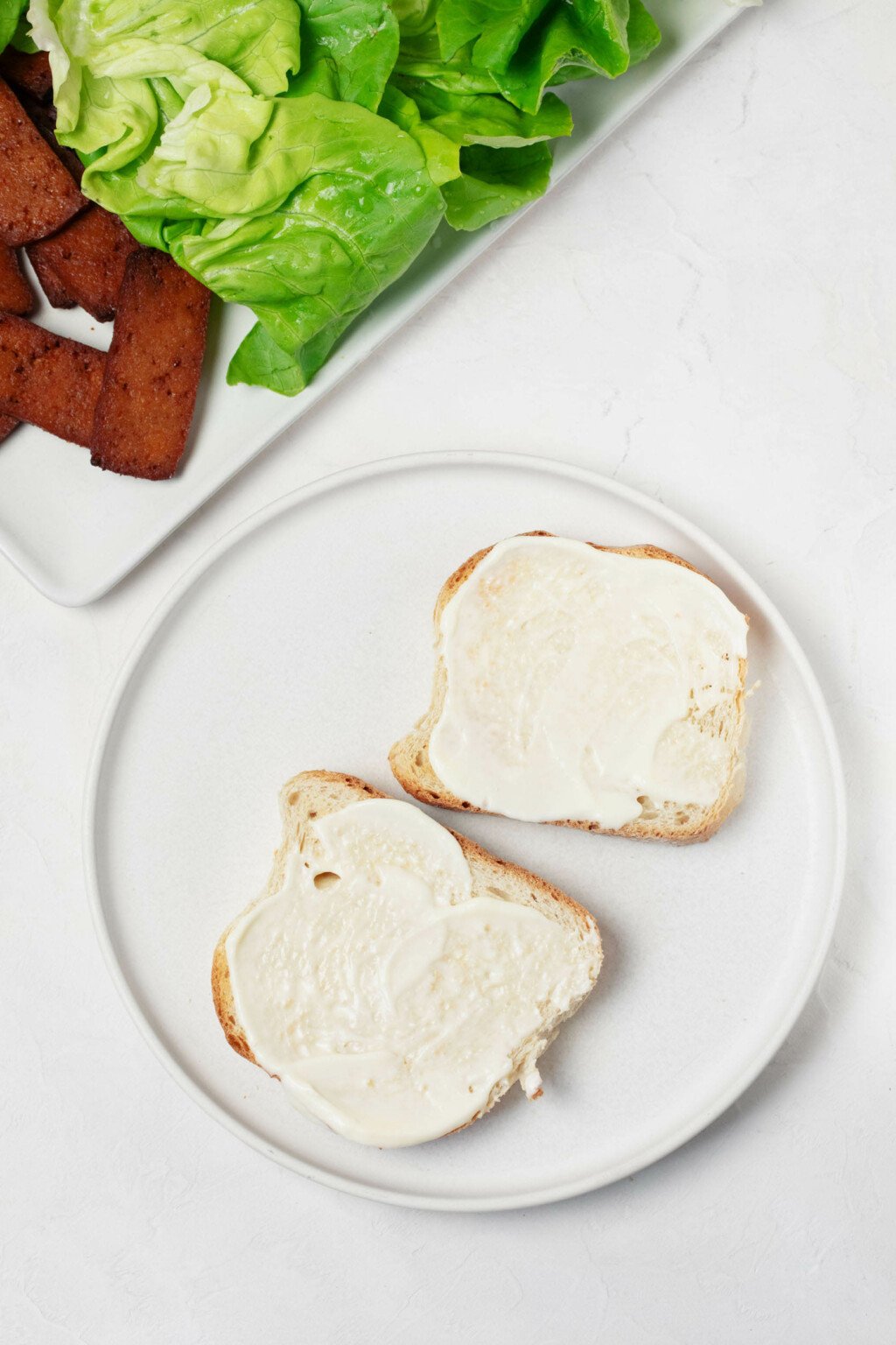 Μια εικόνα από πάνω της ρύθμισης για την προετοιμασία ενός σάντουιτς.  Δύο φέτες ψωμιού επικαλύπτονται με κρεμώδη, κρεμ μαγιονέζα.  Ένας δίσκος με λαχανικά και τόφου είναι κοντά.