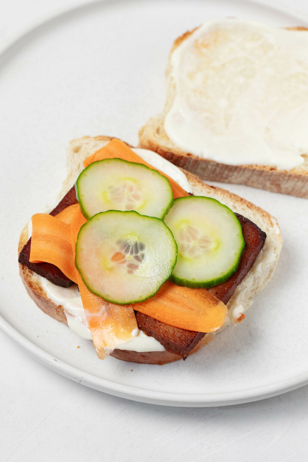 Two slices of toast are visible on a round, rimmed white plate. One slice has pieces of cucumber and carrot, along with baked tofu, layered on top.