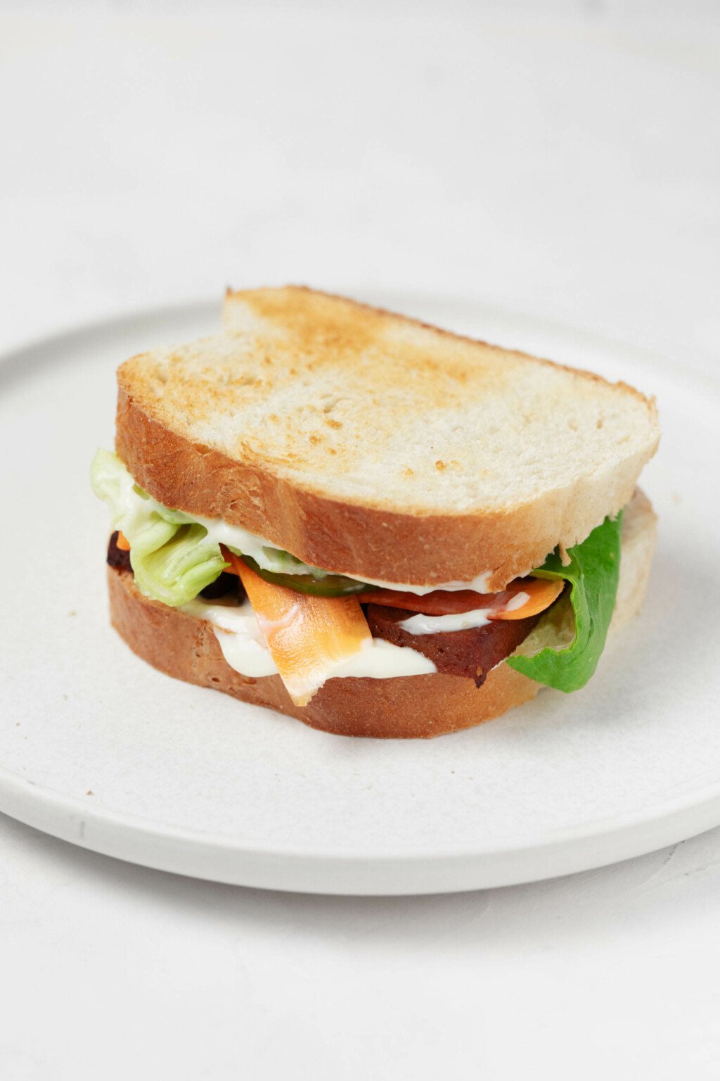 Ένα σάντουιτς, φτιαγμένο με φρυγανισμένο λευκό ψωμί, απεικονίζεται σε ένα στρογγυλό, λευκό πιάτο.  Το σάντουιτς καλύπτεται με μαγιονέζα, λαχανικά τουρσί και φέτες ψημένο τόφου.