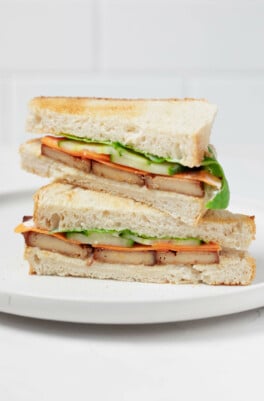 An angled image of a pickled vegetable and smoky tofu sandwich. The sandwich has been cut in half, revealing a cross section with cucumber, carrot, and mayonnaise, along with the tofu.