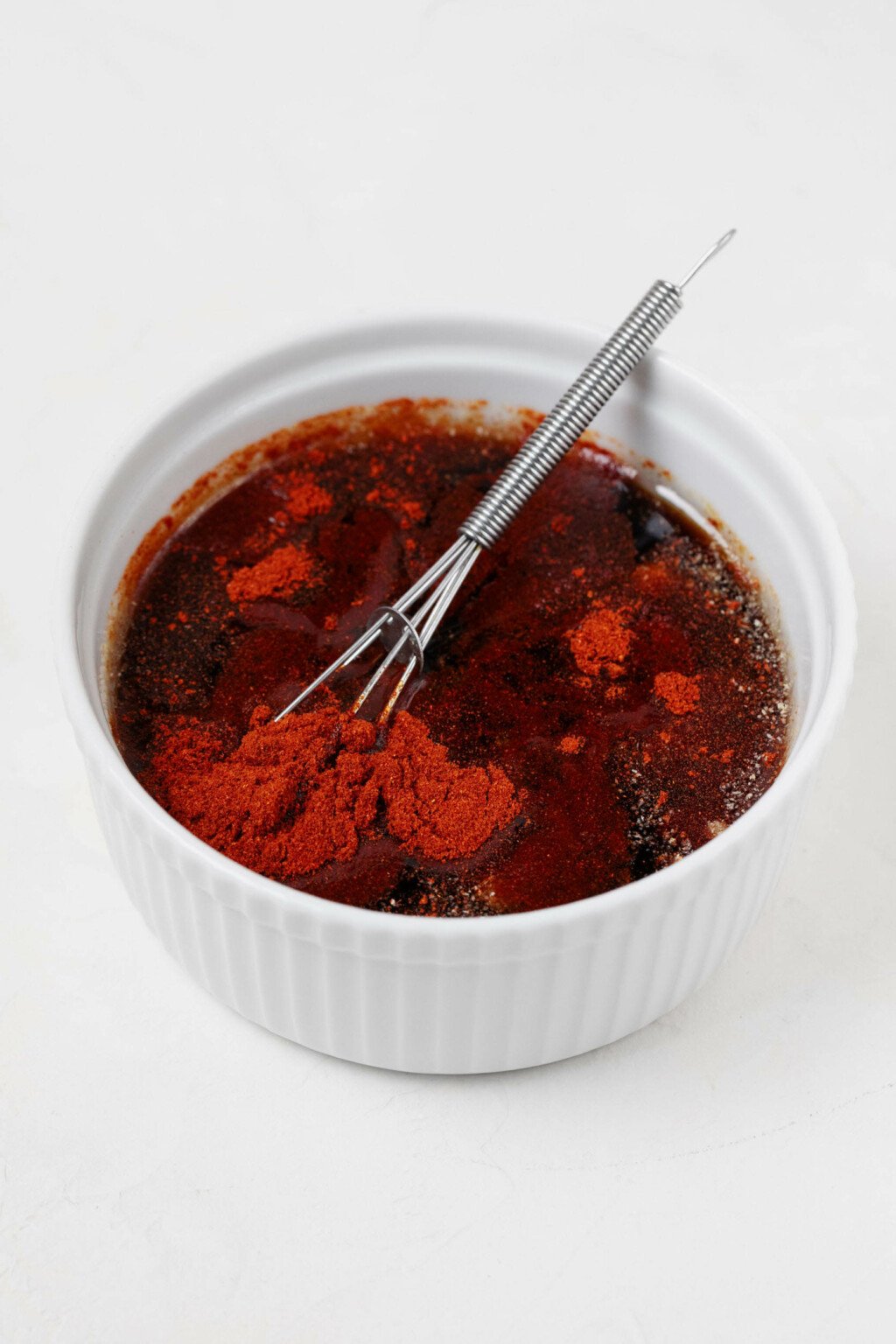 A round, white ramekin is filled with a liquid mixture that's being whisked together. Smoked paprika is visible, turning the marinade a red color.