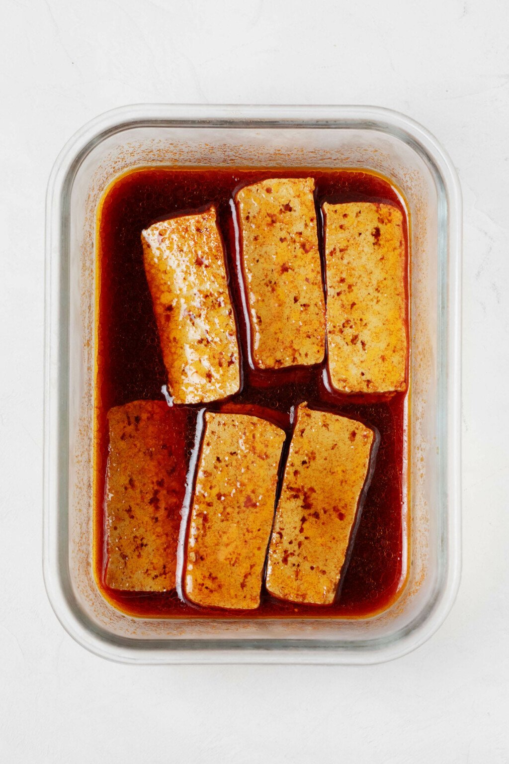 A rectangular storage container holds slices of tofu that are being seasoned with a reddish brown marinade.