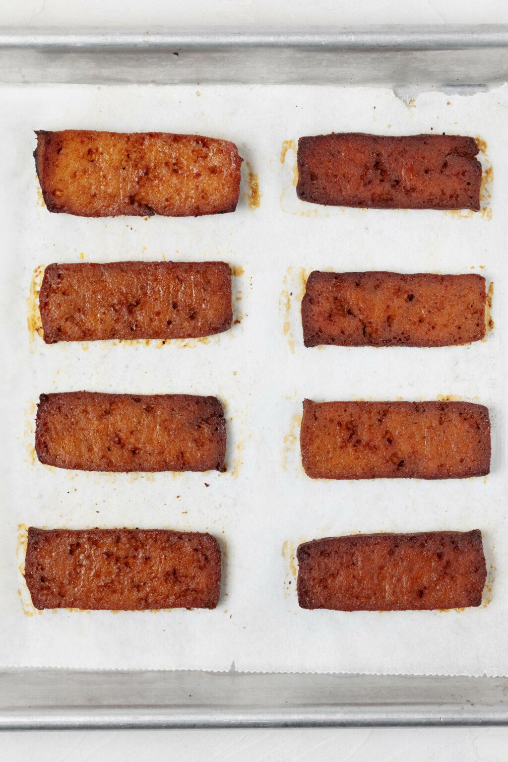 A rectangular, metal baking sheet has been lined with white parchment and sliced, baked tofu slabs.