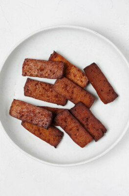 A round, white plate has been covered with rectangular, dark brown slices of baked, smoky tofu.