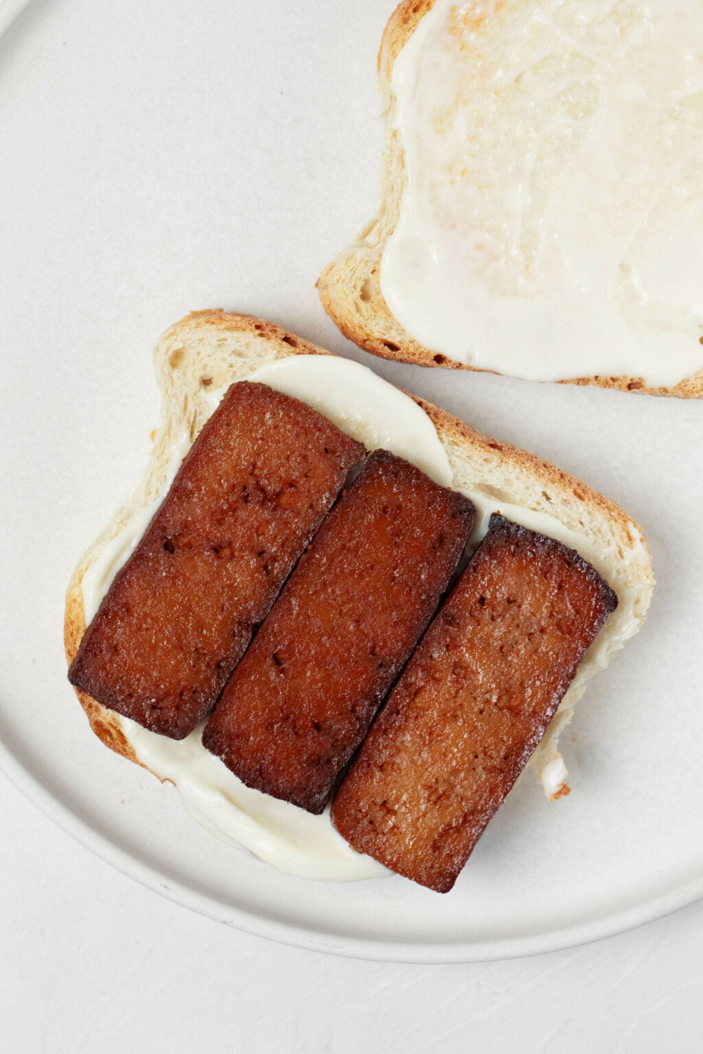A slice of bread is topped with mayonnaise and dark brown slices of smoky tofu.