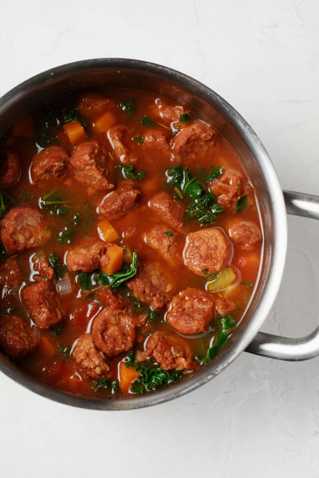 An overhead image of a soup pot, which is filled with a rustic mixture of vegetables, tomato, broth, and vegan sausage slices.