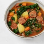 A gray ceramic round bowl has been filled with a hearty plant-based soup. The soup is chock full of kale, potatoes, and vegan sausage slices.