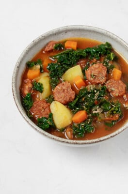 A gray ceramic round bowl has been filled with a hearty plant-based soup. The soup is chock full of kale, potatoes, and vegan sausage slices.