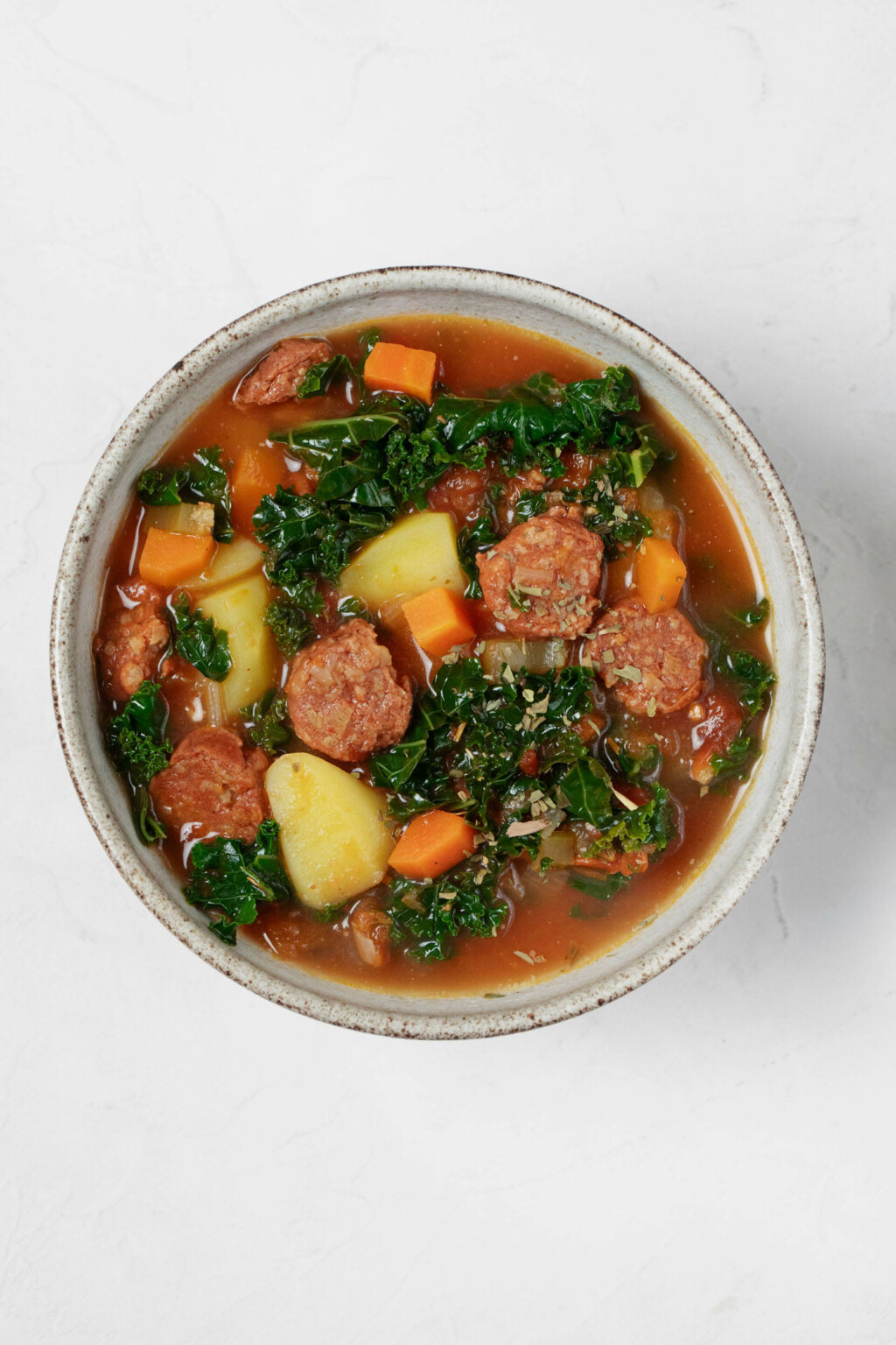 A gray ceramic round bowl has been filled with a hearty plant-based soup. The soup is chock full of kale, potatoes, and vegan sausage slices.