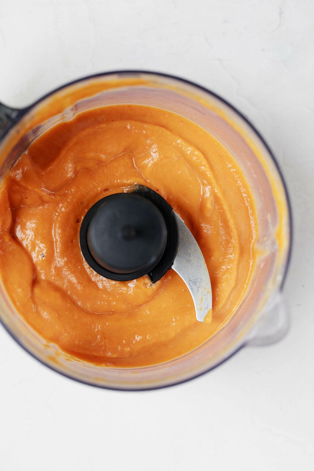 The bowl of a food processor is filled with a pink and orange hued sauce with a creamy consistency.