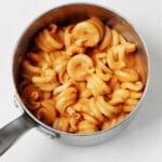A small, silver sauce pan is filled with a creamy pumpkin pasta with an orange hue. It rests on a white surface.