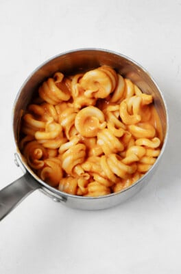 A small, silver sauce pan is filled with a creamy pumpkin pasta with an orange hue. It rests on a white surface.