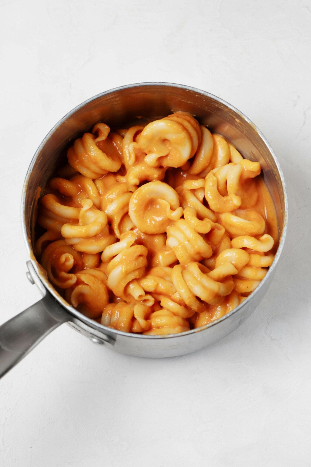 A small, silver sauce pan is filled with a creamy pumpkin pasta with an orange hue. It rests on a white surface.
