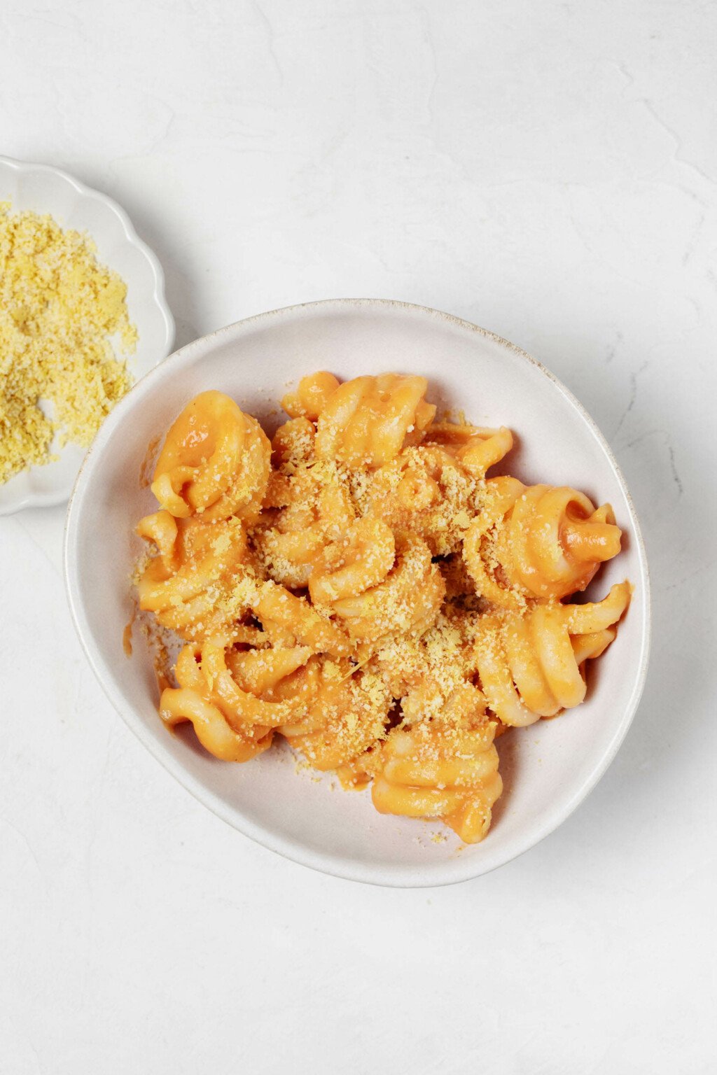 An overhead image of a small, white bowl filled with a creamy vegan pumpkin pasta. A pinch bowl of vegan parmesan cheese rests nearby.