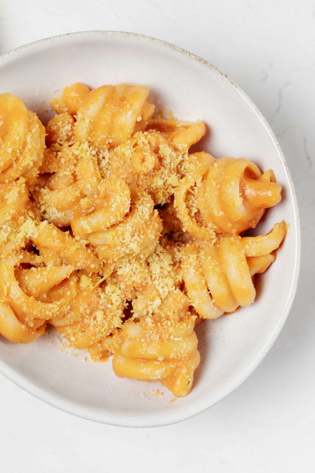 An overhead image of a bowl of creamy plant-based pumpkin pasta. The pasta is topped with a homemade cashew "parmesan" cheese. The bowl rests on a white surface.