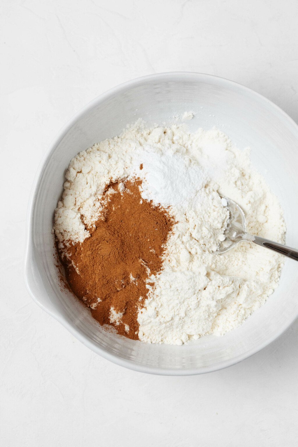 A mixture of flour and baking spices is pictured in a large, white mixing bowl. A whisk is standing in the bowl. 