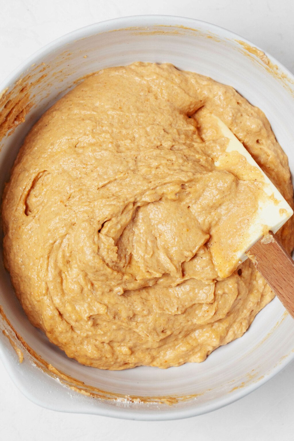 A thick, orange-hued waffle batter is being mixed in a large white mixing bowl with a spatula.