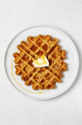A round, white plate rests on a white surface. The plate is being used to serve a vegan pumpkin waffle, which is topped with plant-based butter and maple syrup.
