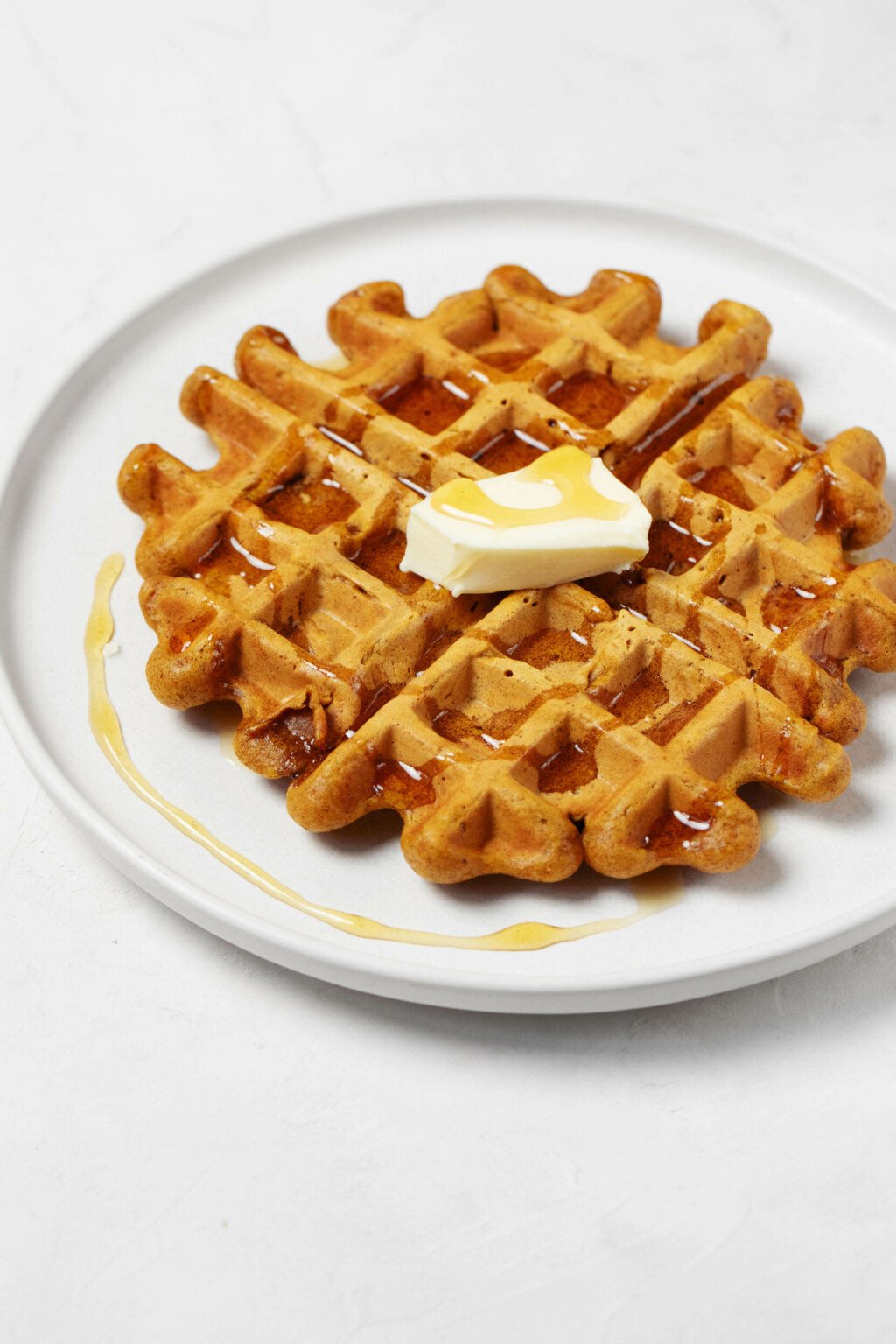 A round, white plate is topped with a deeply golden colored, toasted waffle. The waffle has been topped with butter and syrup.