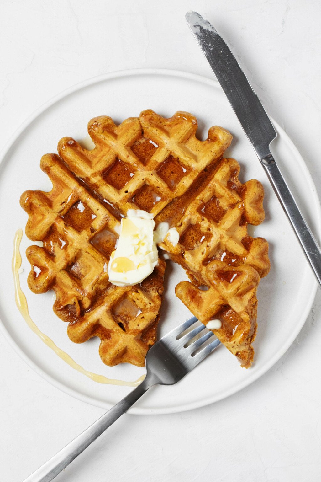 An overhead image of a vegan pumpkin waffle, which is being served on a round, white plate. A fork and knife are being used to slice the waffle, which is topped with a pat of butter and a drizzle of maple syrup.