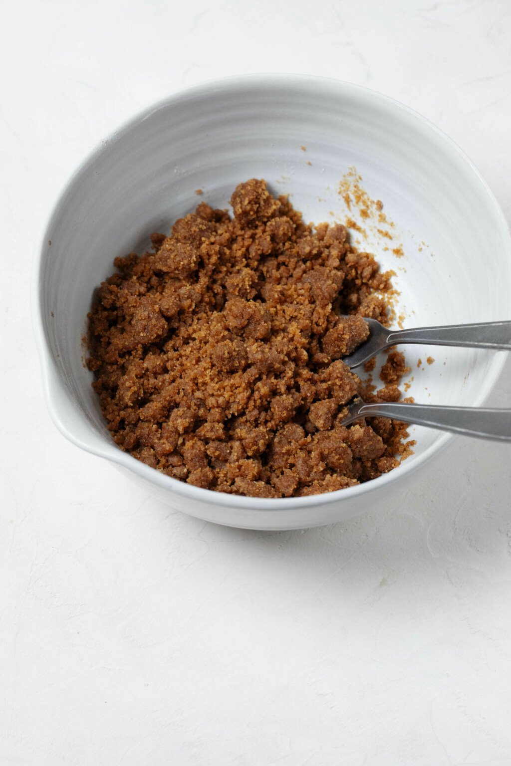 A white, ceramic mixing bowl is filled with a vegan streusel topping. The topping is made with dark brown sugar and has big crumbs. Two forks peek out of the bowl.
