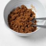 A white, ceramic mixing bowl is filled with a vegan streusel topping. The topping is made with dark brown sugar and has big crumbs. Two forks peek out of the bowl.