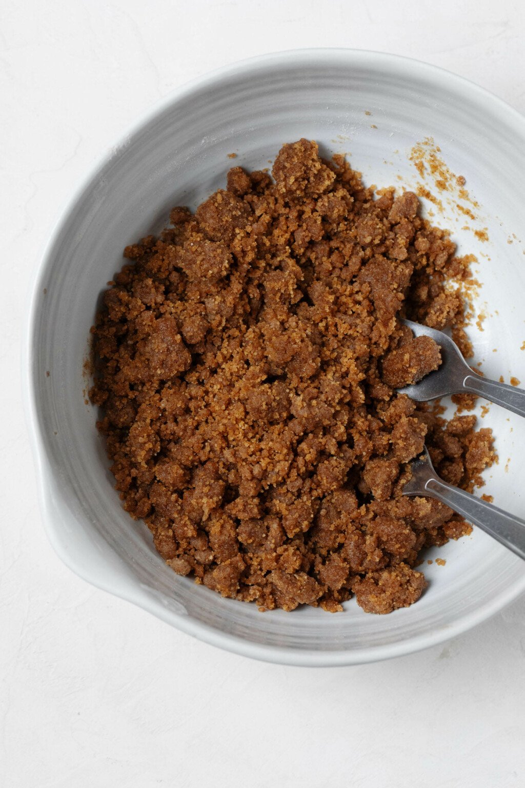 A white mixing bowl is being used to hold a plant-based streusel topping. There are big, deep brown crumbs pictured in the bowl.
