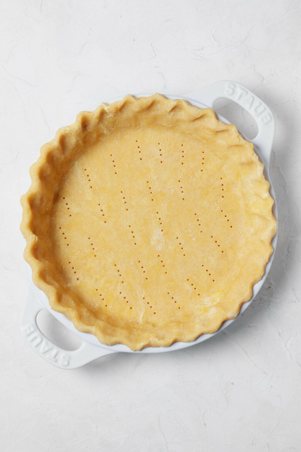An overhead image of a pie crust, which has been arranged in a white, ceramic pie plate. It rests on a white surface.