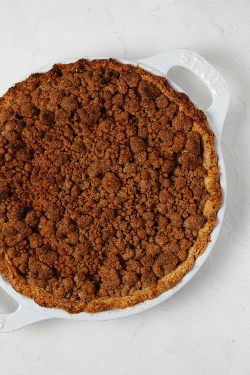 An overhead image of a vegan apple pie with streusel topping, which has just emerged from the oven in a golden brown state. The pie is in a white ceramic pie plate.