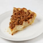 A hearty slice of vegan apple pie with streusel topping has been arranged on a small, round white plate.