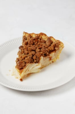 A hearty slice of vegan apple pie with streusel topping has been arranged on a small, round white plate.