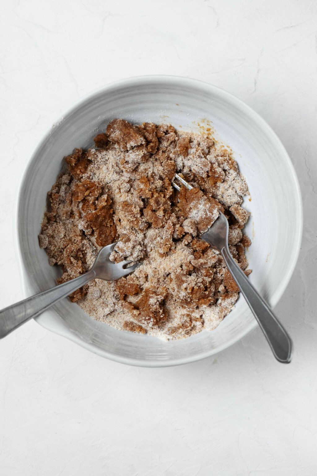A white mixing bowl is pictured overhead, resting on a white surface. There are two forks in the bowl, which are being used to mix a vegan streusel topping.