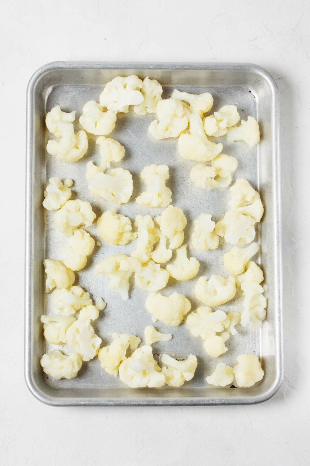 An overhead image of a metal baking sheet, which has been covered with frozen florets of white cauliflower. 