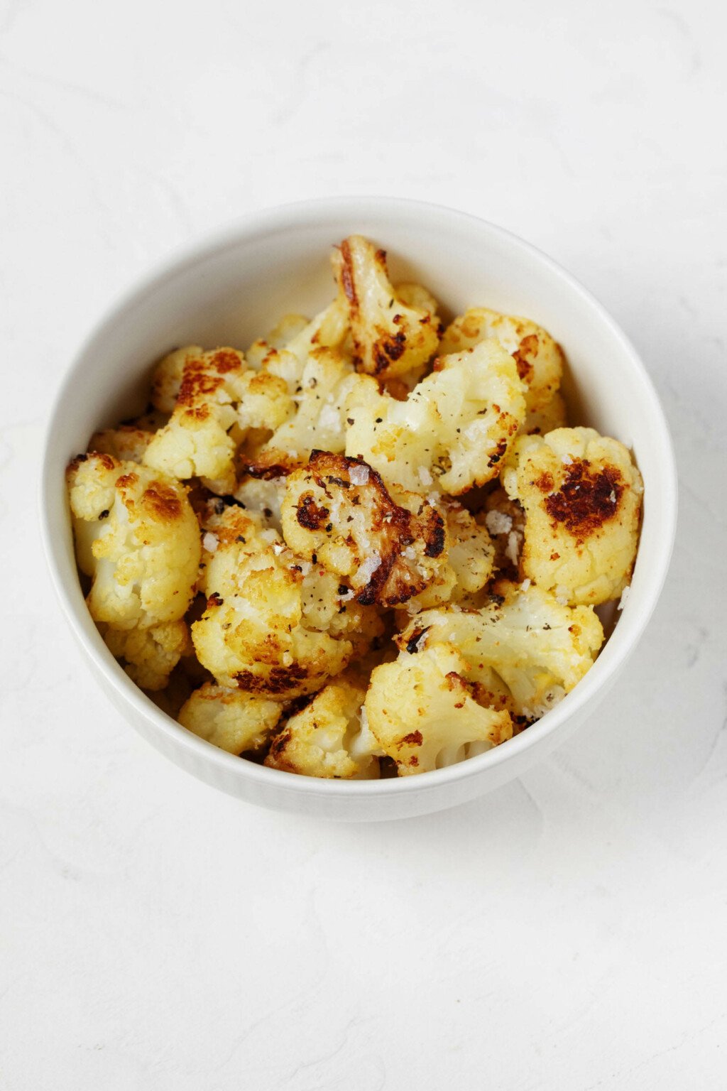 An overhead image of a white bowl, which is filled with roasted frozen cauliflower florets that are topped with flaky sea salt.