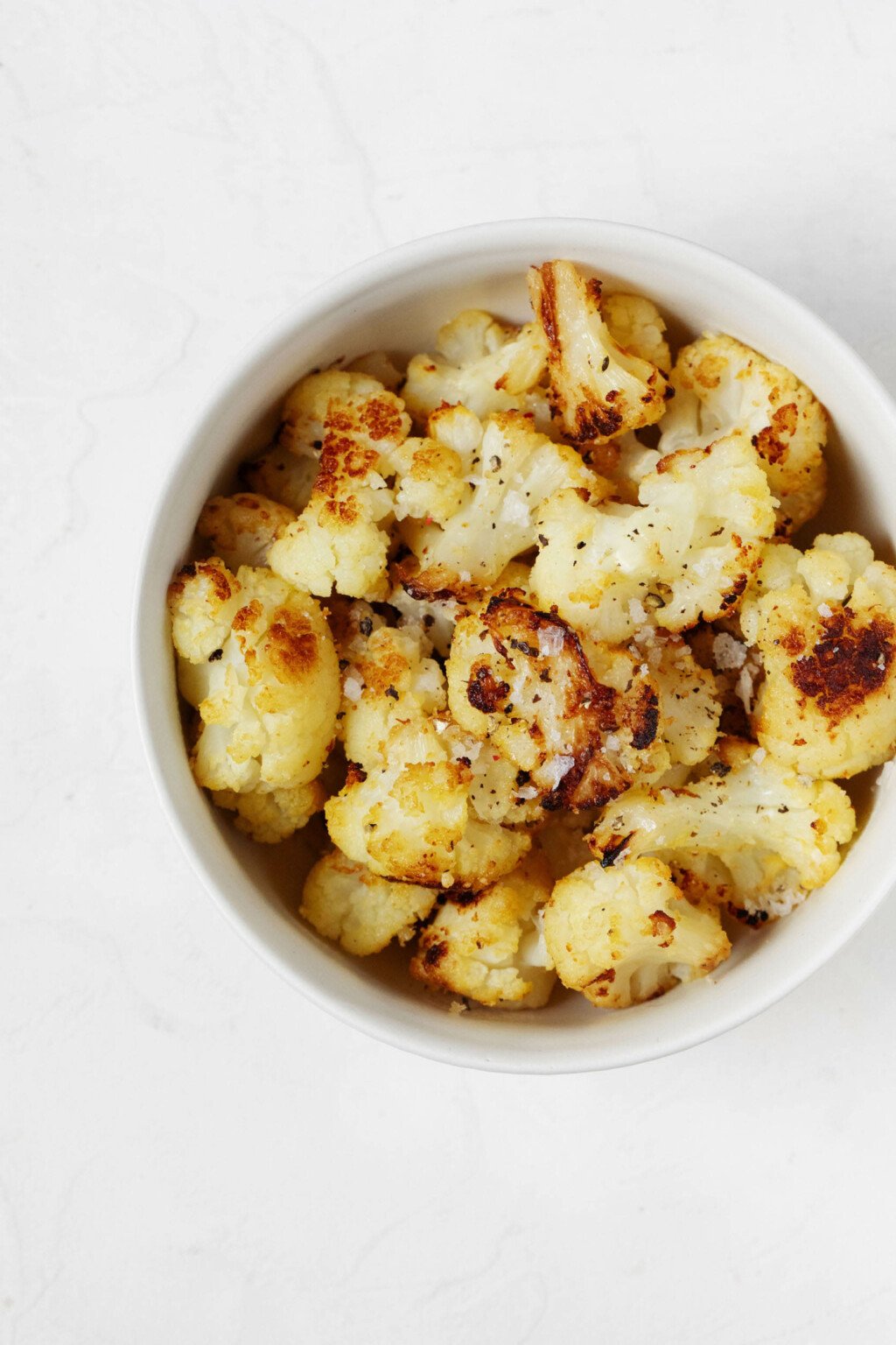 An overhead image of a white bowl, which is filled with roasted frozen cauliflower florets that are topped with flaky sea salt.