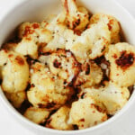 An overhead image of a white bowl, which is filled with roasted frozen cauliflower florets that are topped with flaky sea salt.